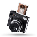 A 3/4 angle view of a black instax camera with silver details and a portrait photo in the eject slot on top.