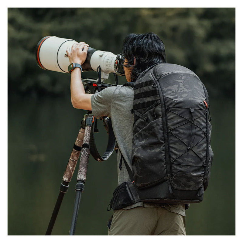 Outdoor photographer wearing PGYTECH OnePro ST Backpack with camouflage pattern using camera and tripod by water